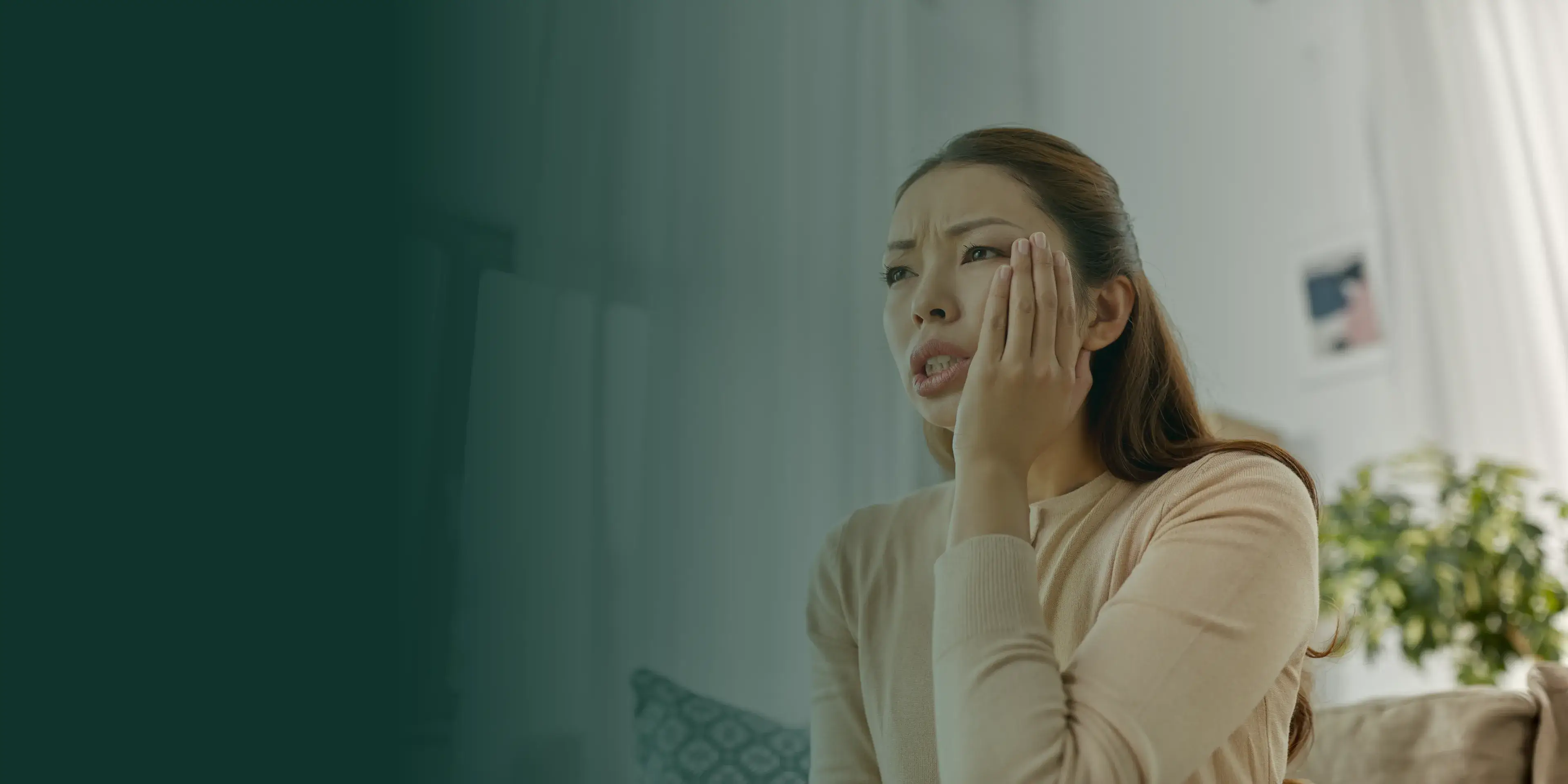 A woman holds her jaw in pain, sitting on a couch at home.