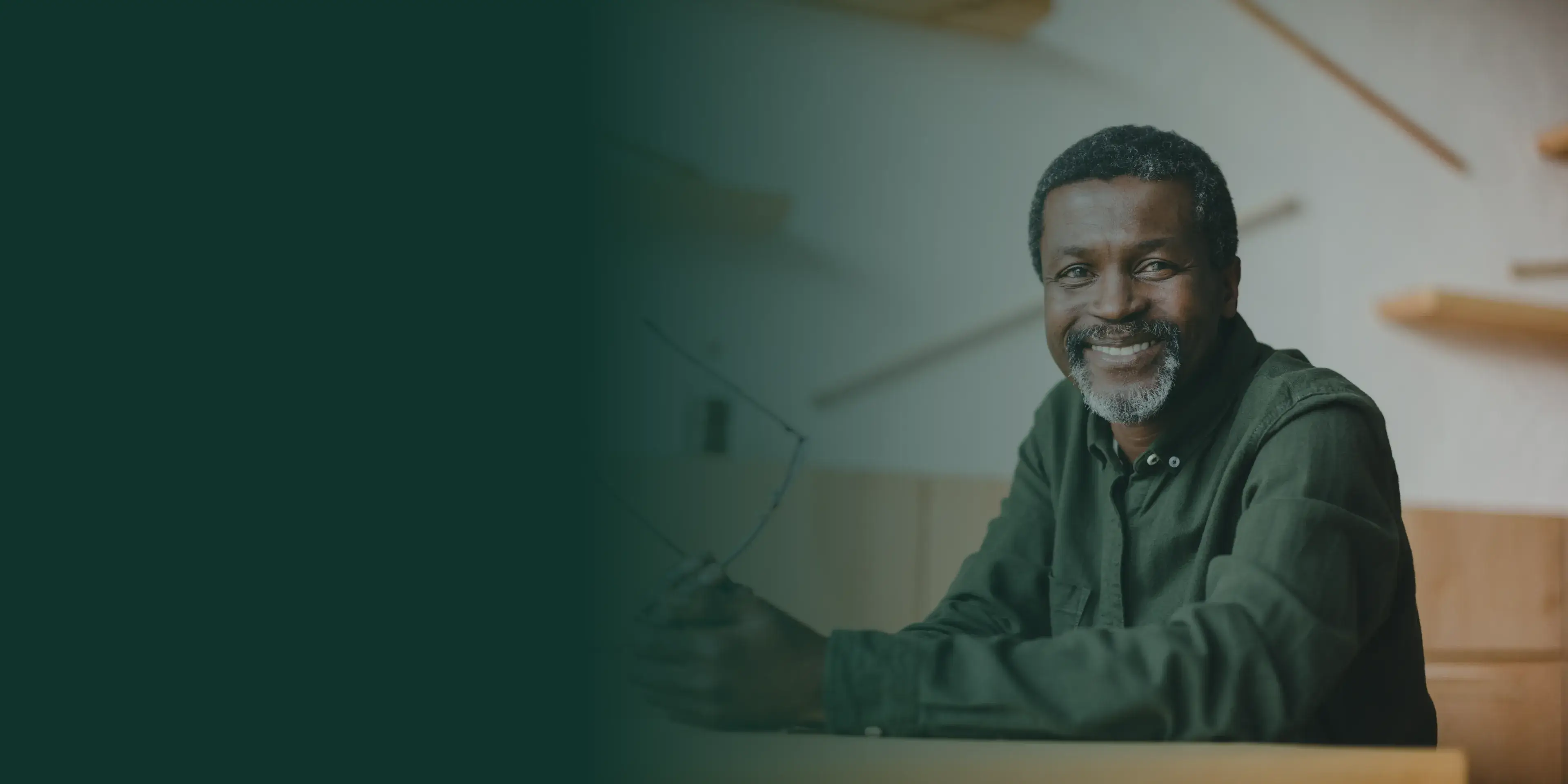 Smiling man in a green shirt sits indoors, holding glasses and looking toward the camera.
