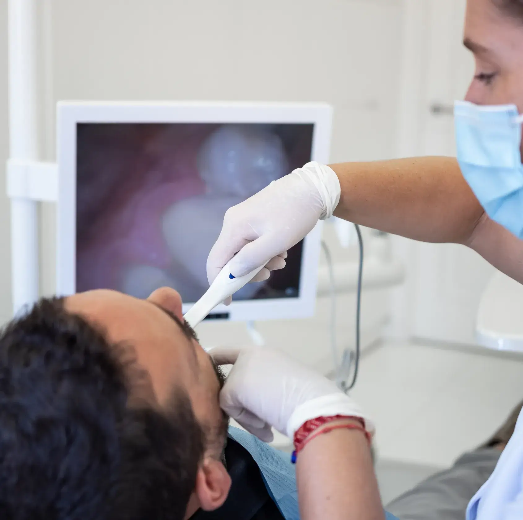 A dentist examines a patient's mouth with a dental instrument, while a screen shows an image of teeth.