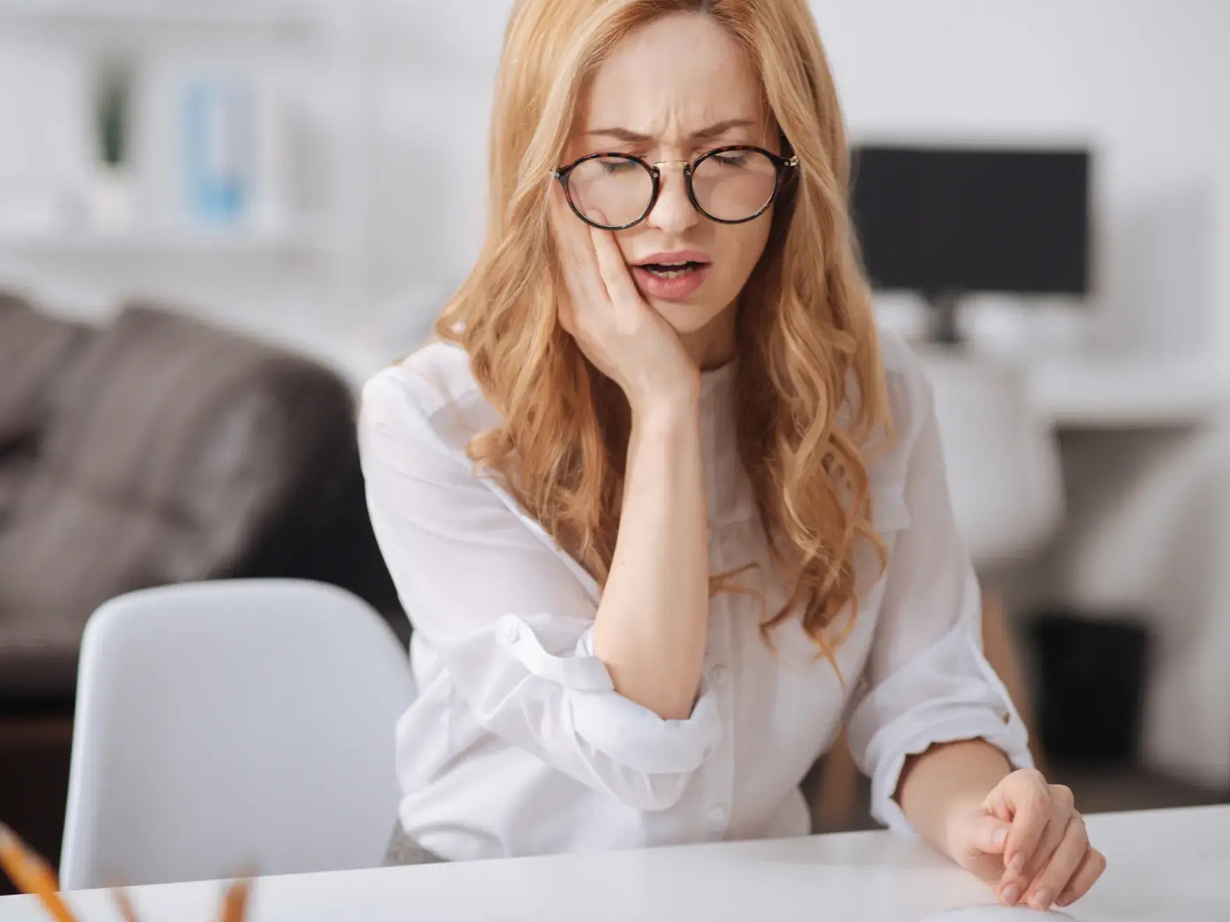 Woman in glasses sitting at a table, holding her cheek with an expression of discomfort.