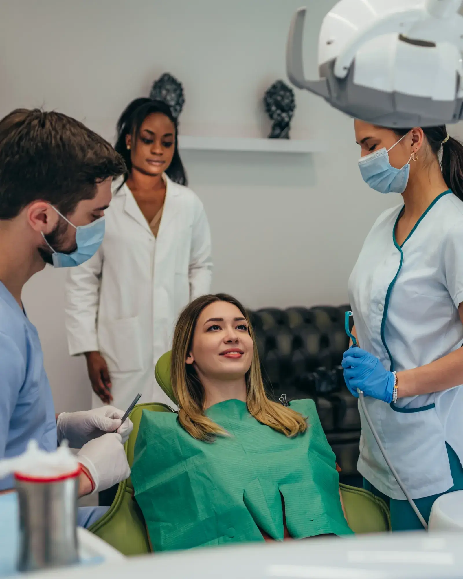 A woman sits in a dental chair while two dental professionals and an assistant stand nearby.