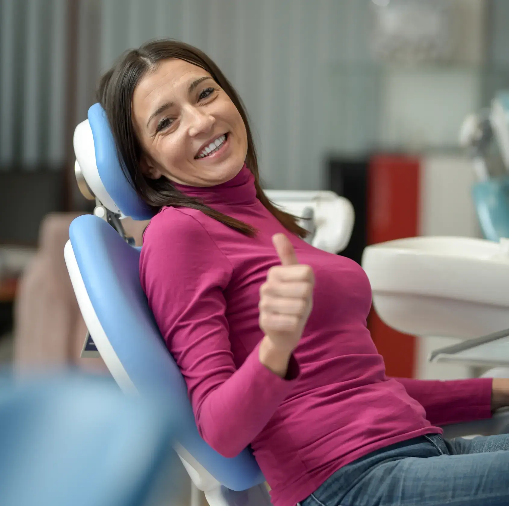 A woman in a pink shirt sits in a dentist chair, smiling and giving a thumbs-up.