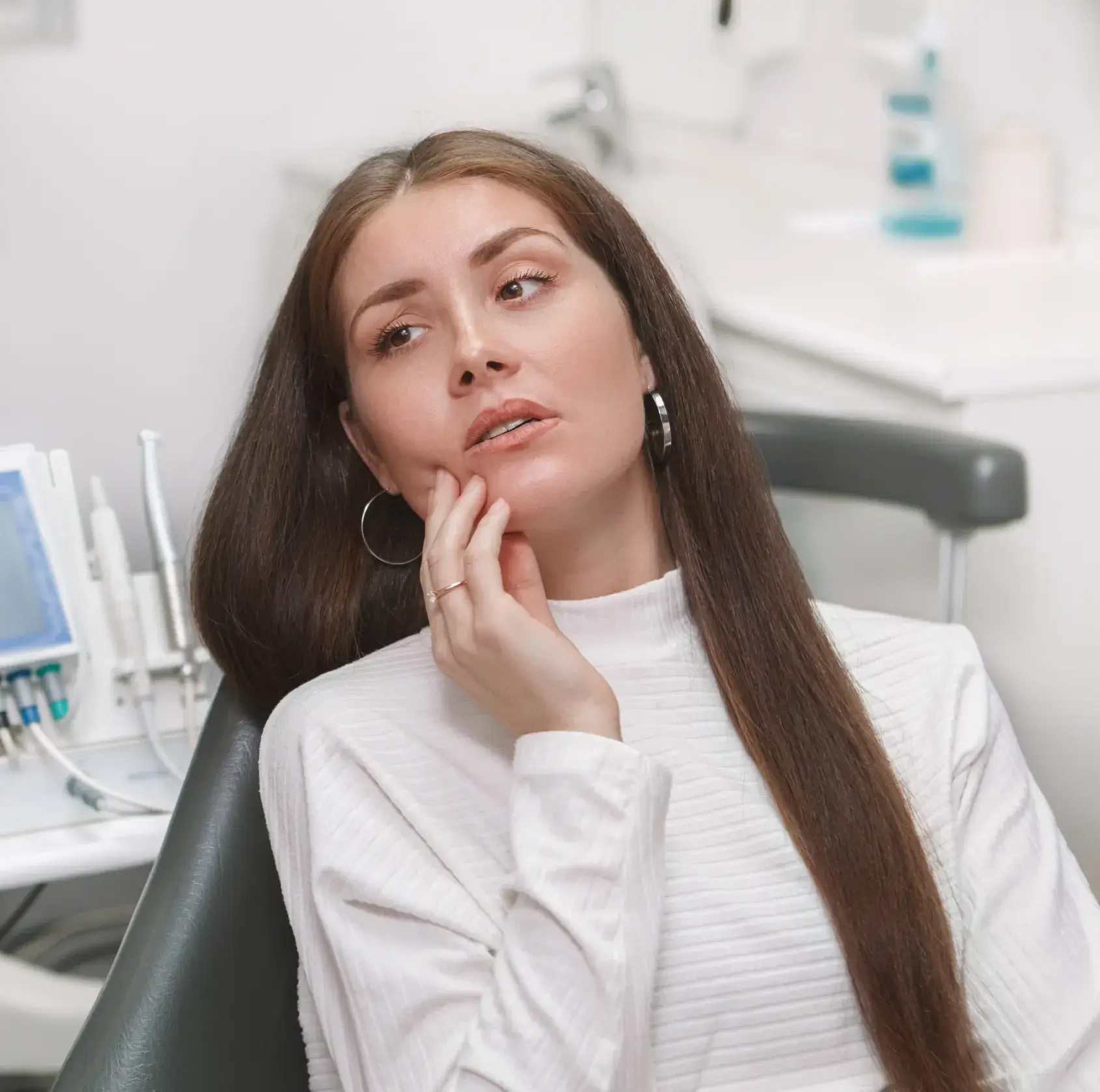 Woman in a dentist chair touching her cheek, looking concerned.