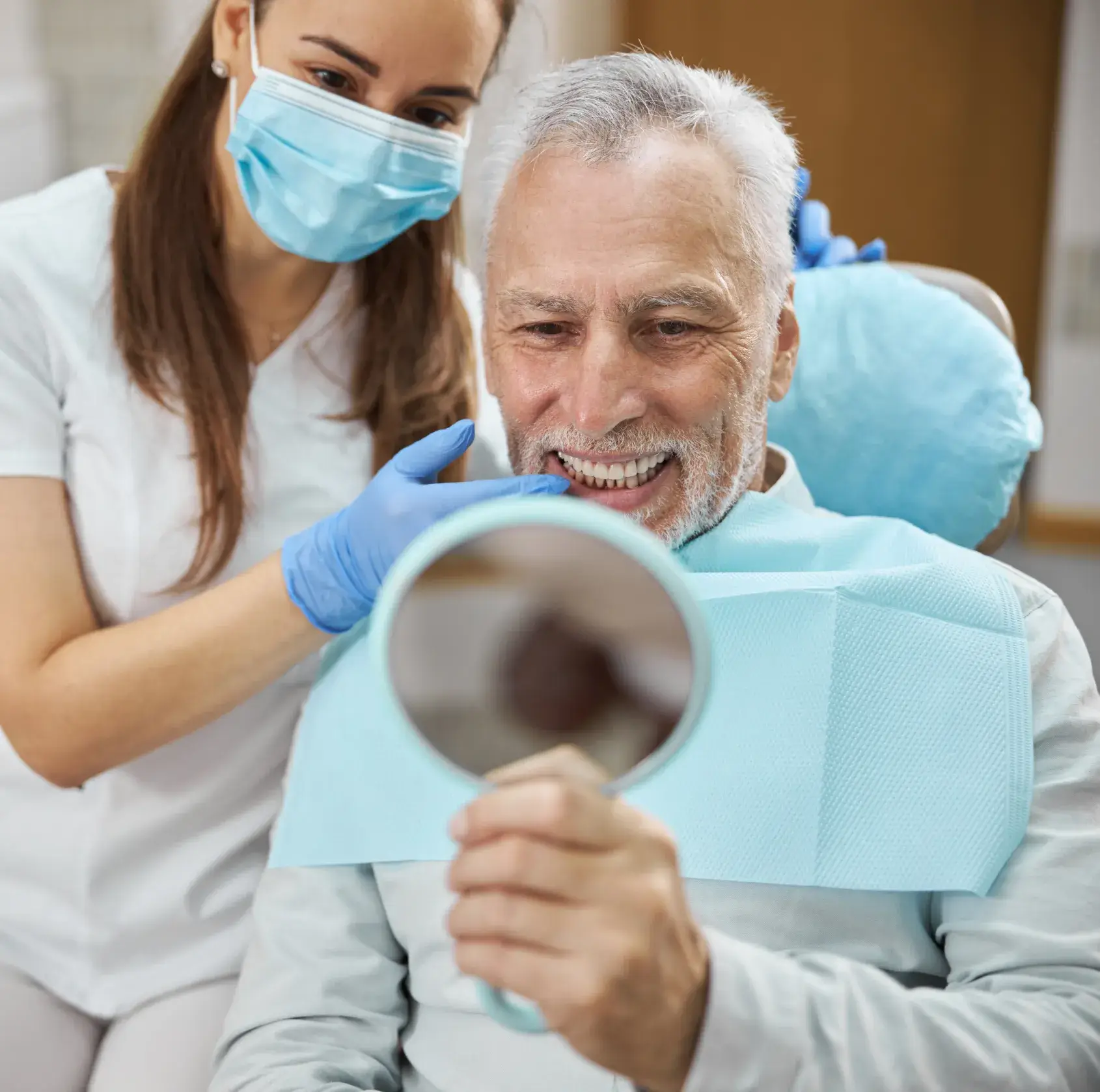 A dentist in blue gloves shows a smiling man his teeth in a small mirror.