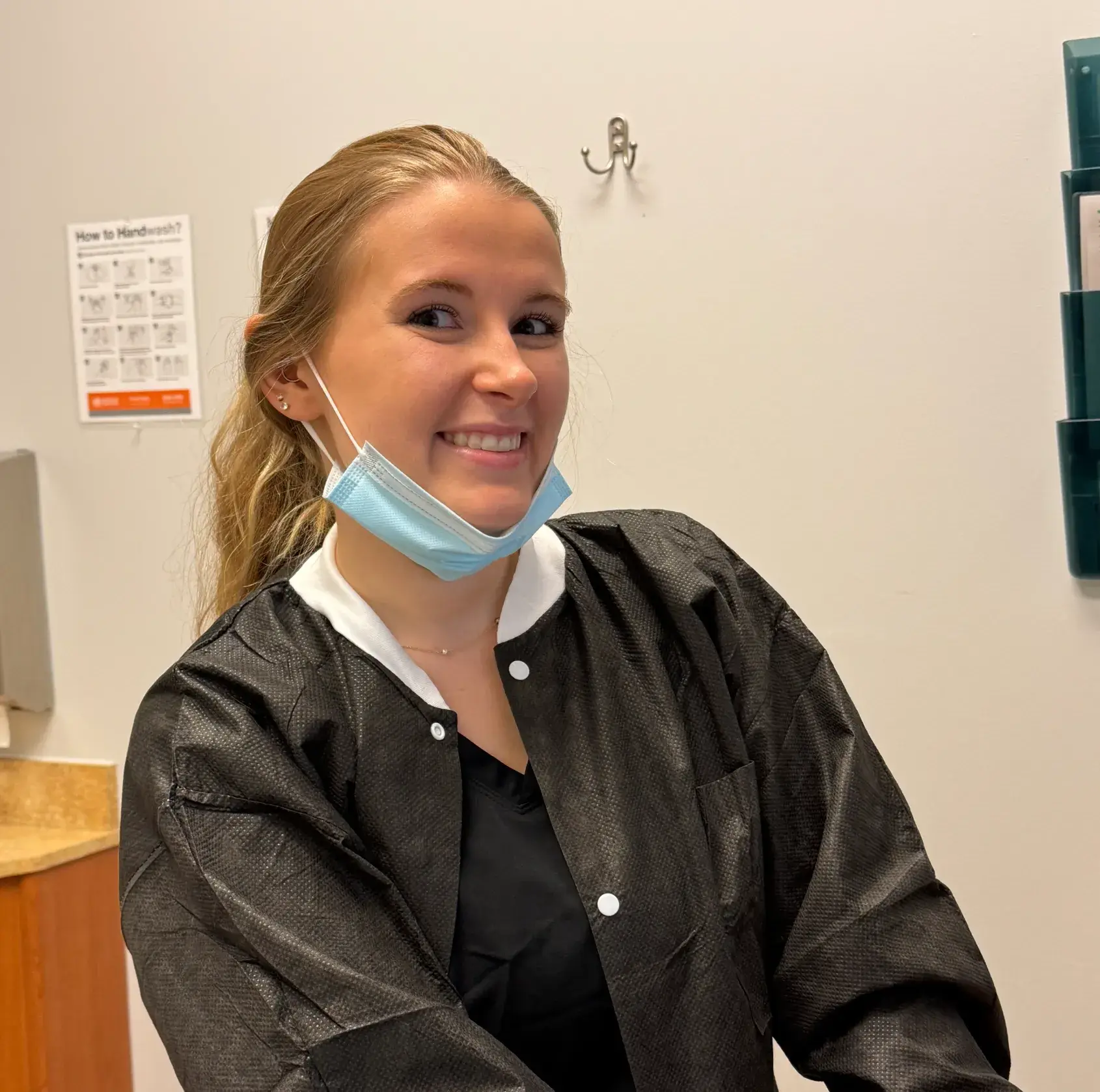 A woman wearing a black medical uniform smiles and poses with a mask under her chin.