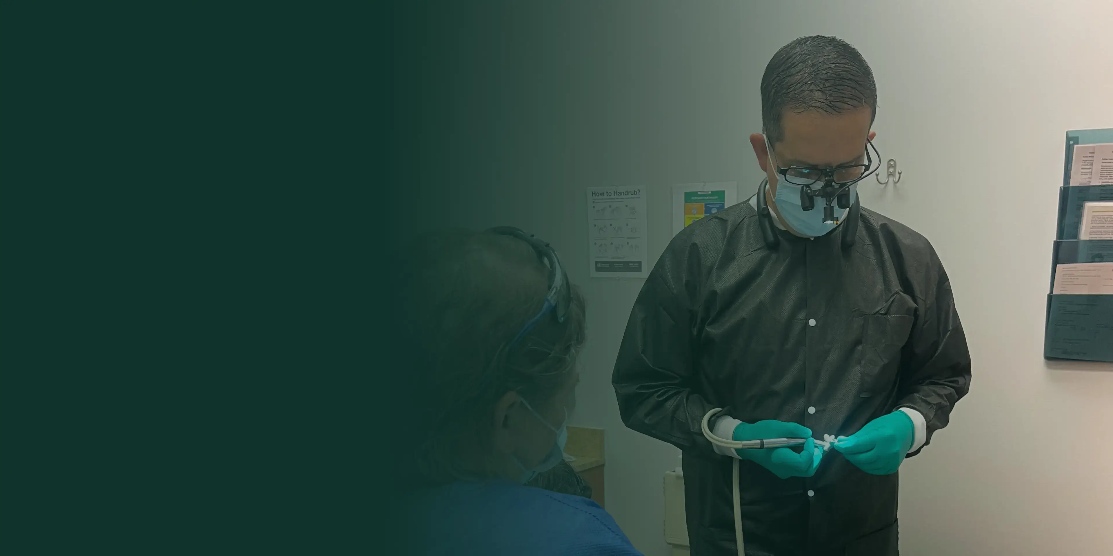 Dentist wearing magnifying glasses and mask examines a dental tool while a patient sits nearby.