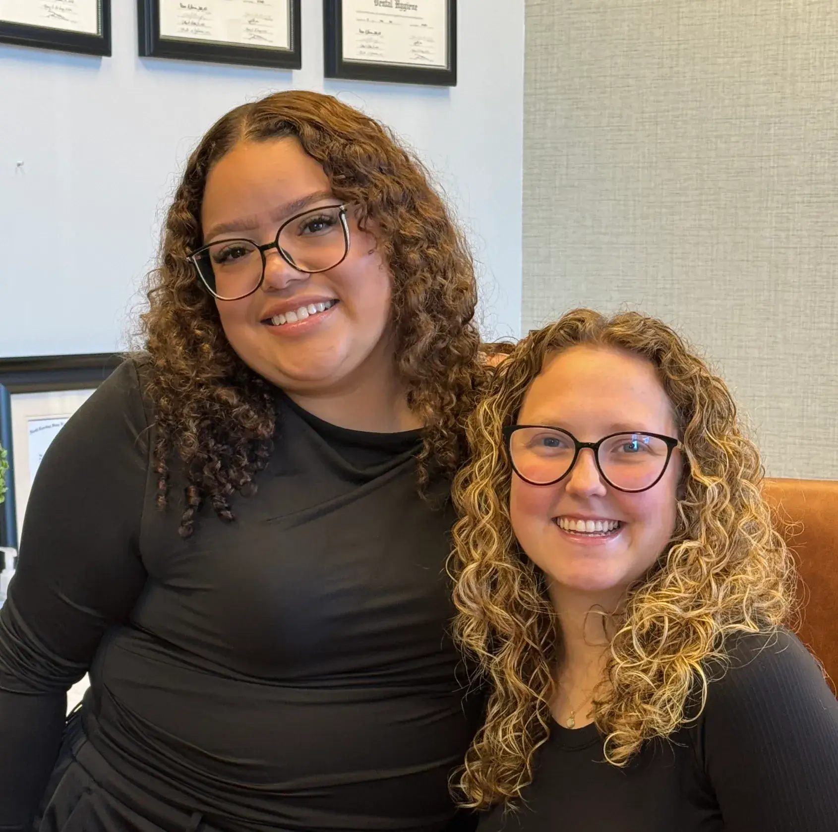 Two women with curly hair and glasses smiling while sitting together in an office setting.