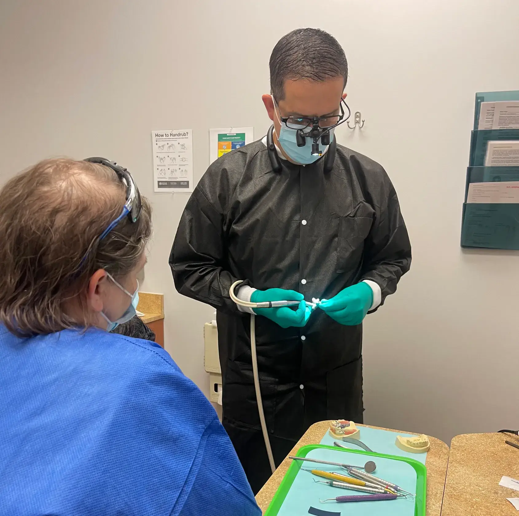 A dentist wearing protective gear examines dental equipment while a patient watches.