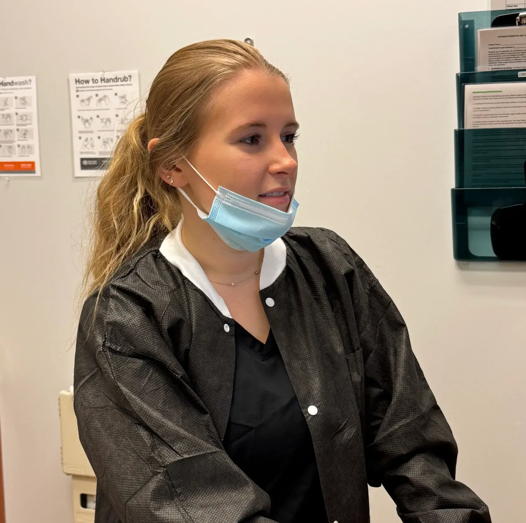 Young woman in a black gown wearing a face mask, standing indoors with medical posters behind her.