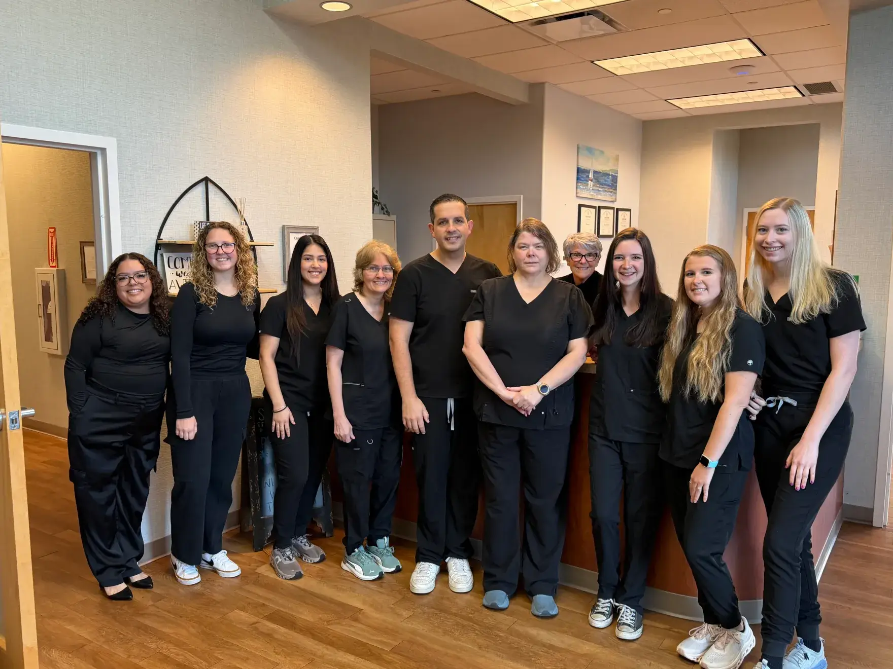 A group of ten people wearing black medical scrubs stand together and smile in an office setting.