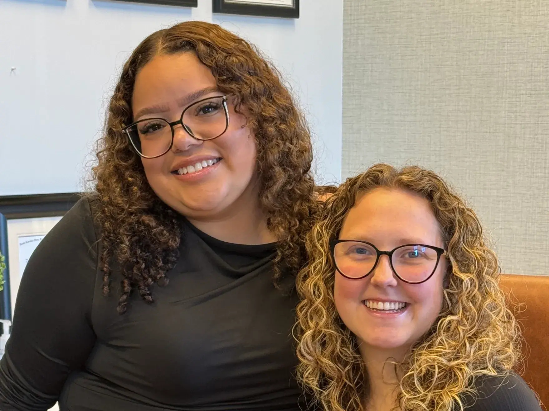 Two people with curly hair and glasses smiling, sitting closely together in a well-lit room.