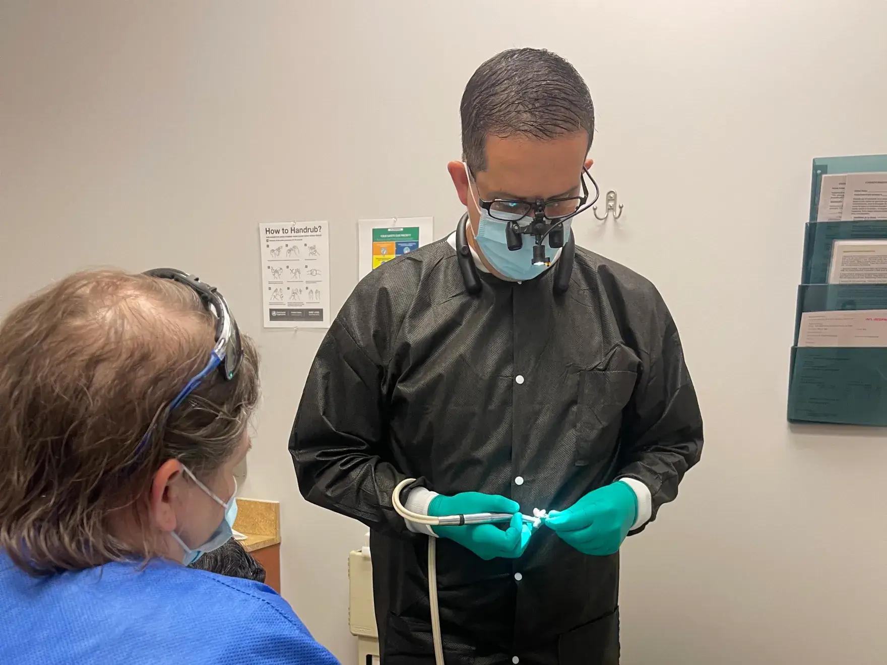 A dentist wearing magnifying glasses prepares dental tools while a patient sits nearby in a clinic.