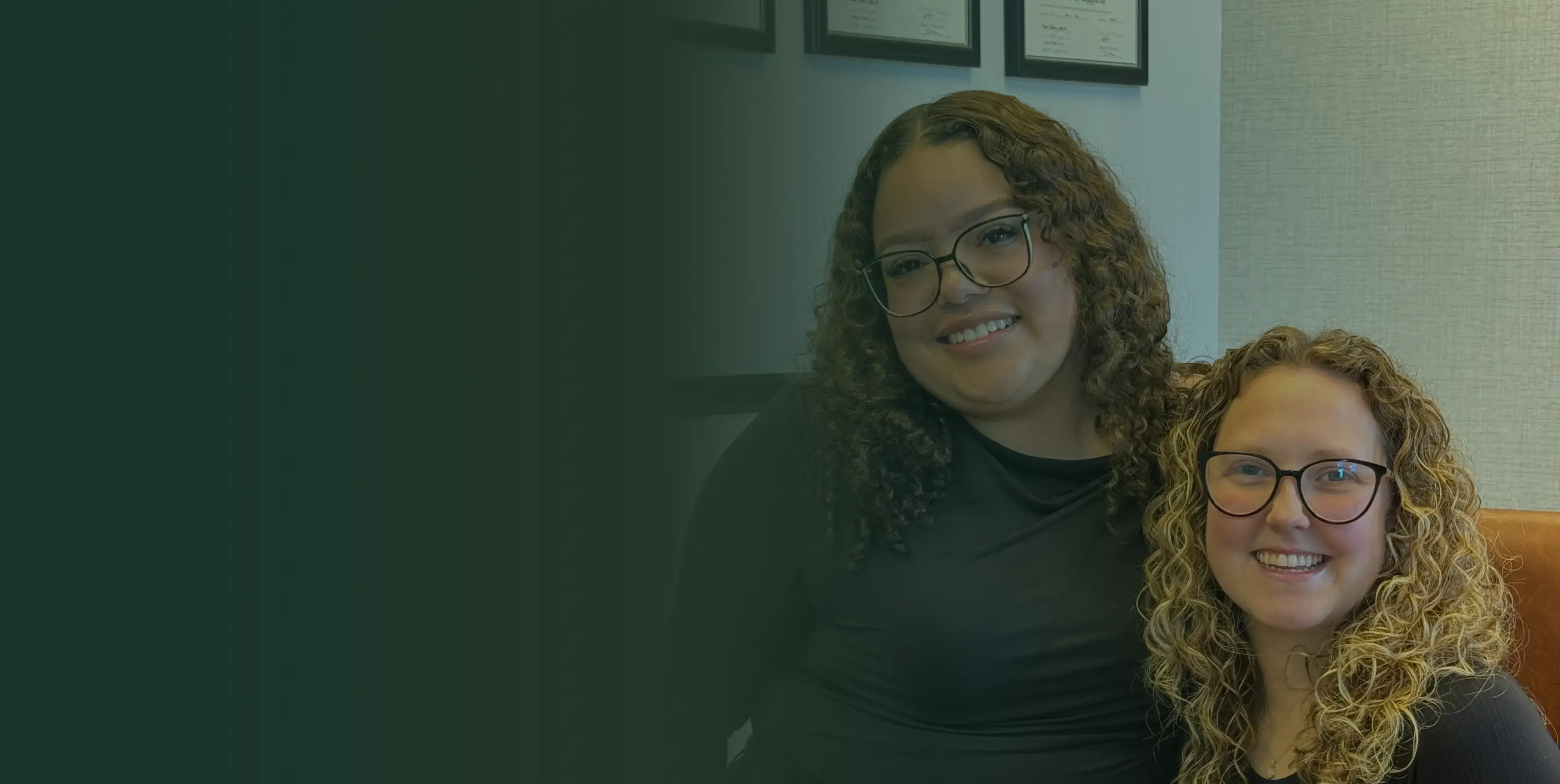 Two people with curly hair, wearing glasses and smiling, sit closely together indoors.