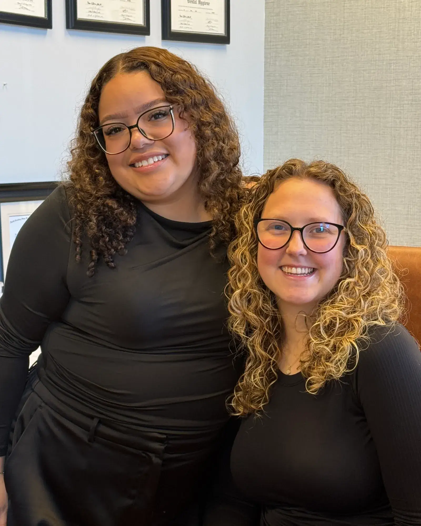 Two people with curly hair and glasses are smiling while posing together indoors.