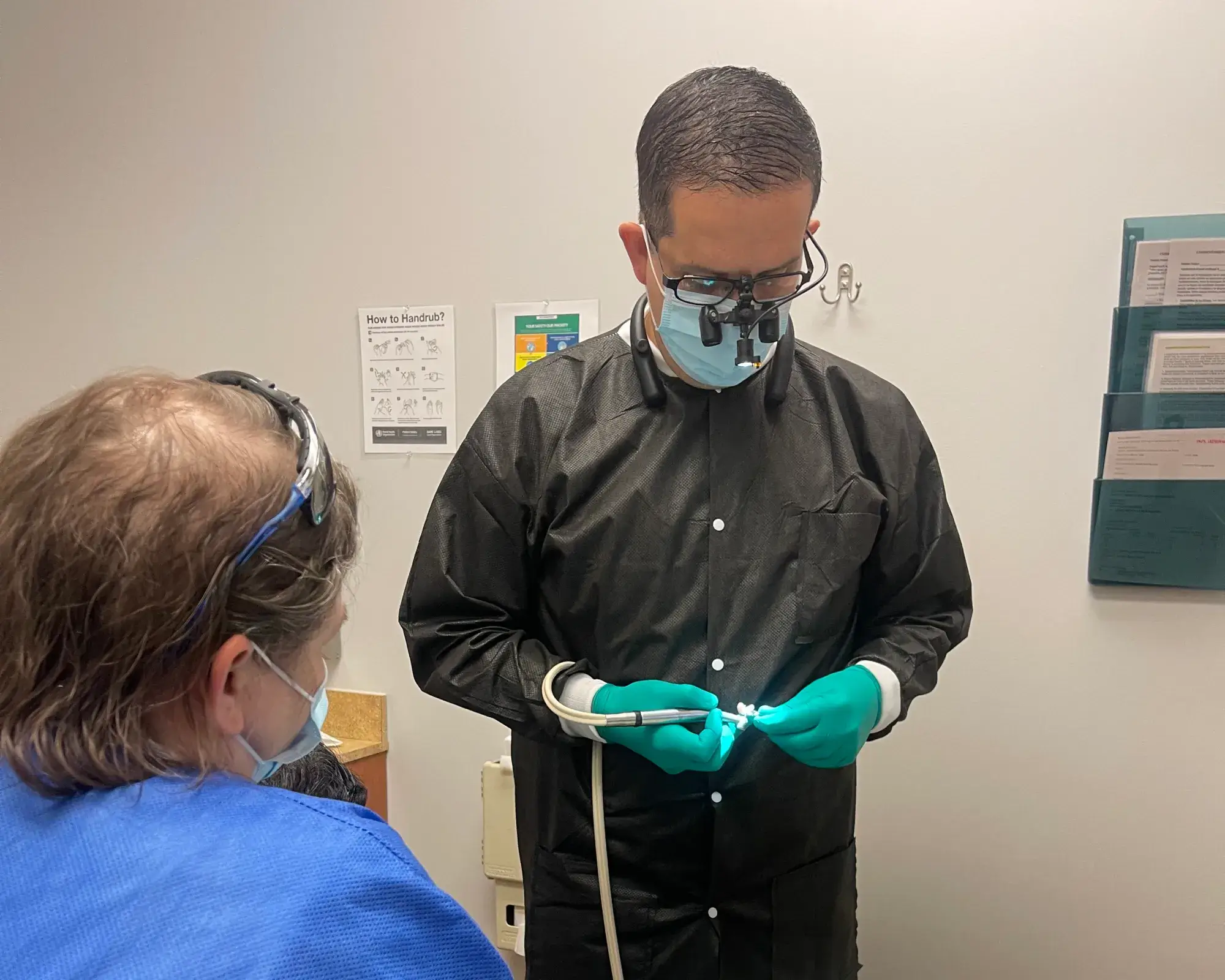 A dentist wearing magnifying glasses examines a dental mold while a patient looks on.
