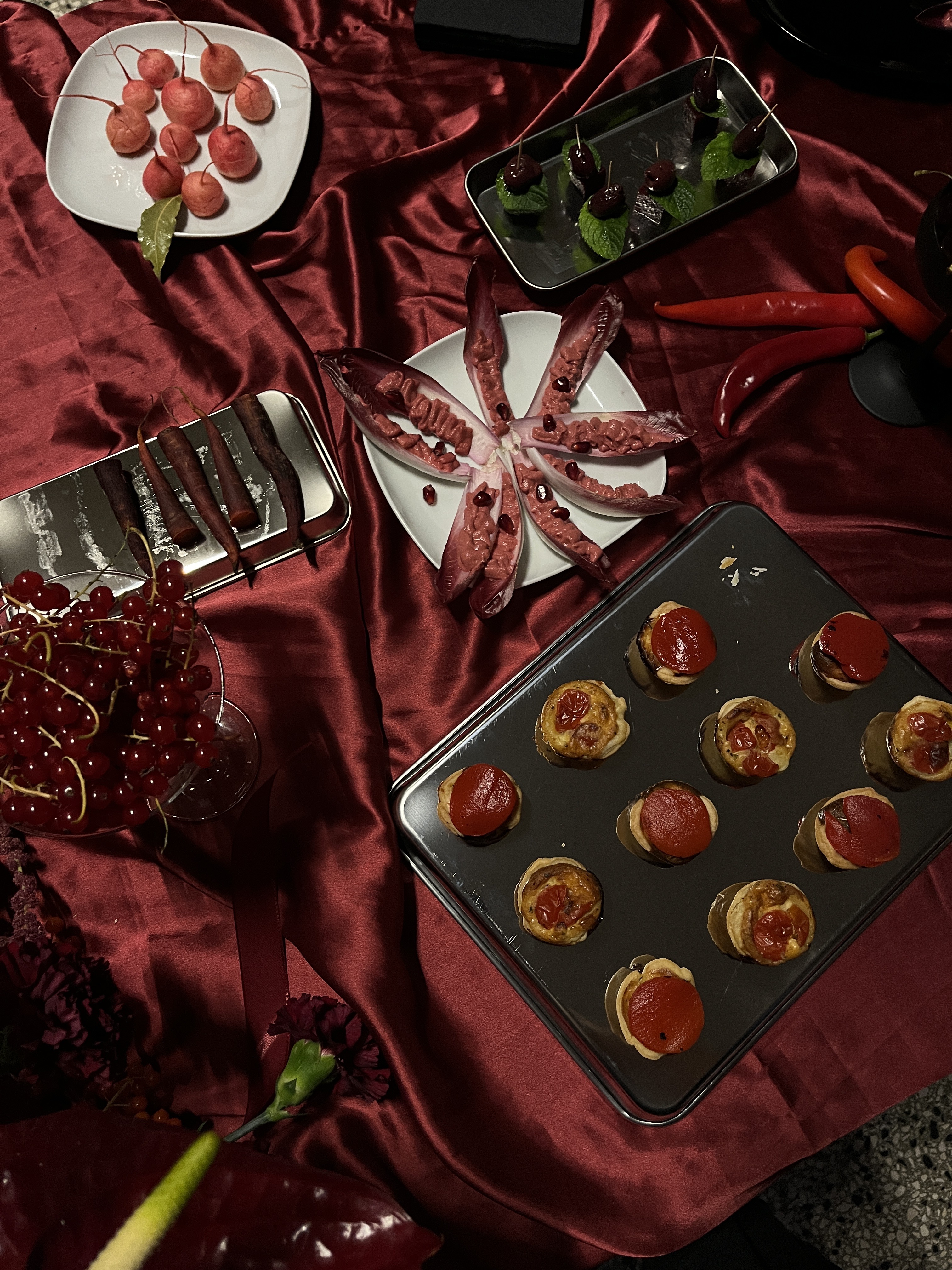Assorted appetizers including mini tarts topped with tomato slices, red endive leaves with spread and pomegranate seeds, skewers with cherries and mint, radishes on a white plate, and red grapes in a glass, arranged on a red satin tablecloth.