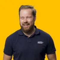 Smiling man with a beard and short hair wearing a navy blue polo shirt standing against a bright yellow background.
