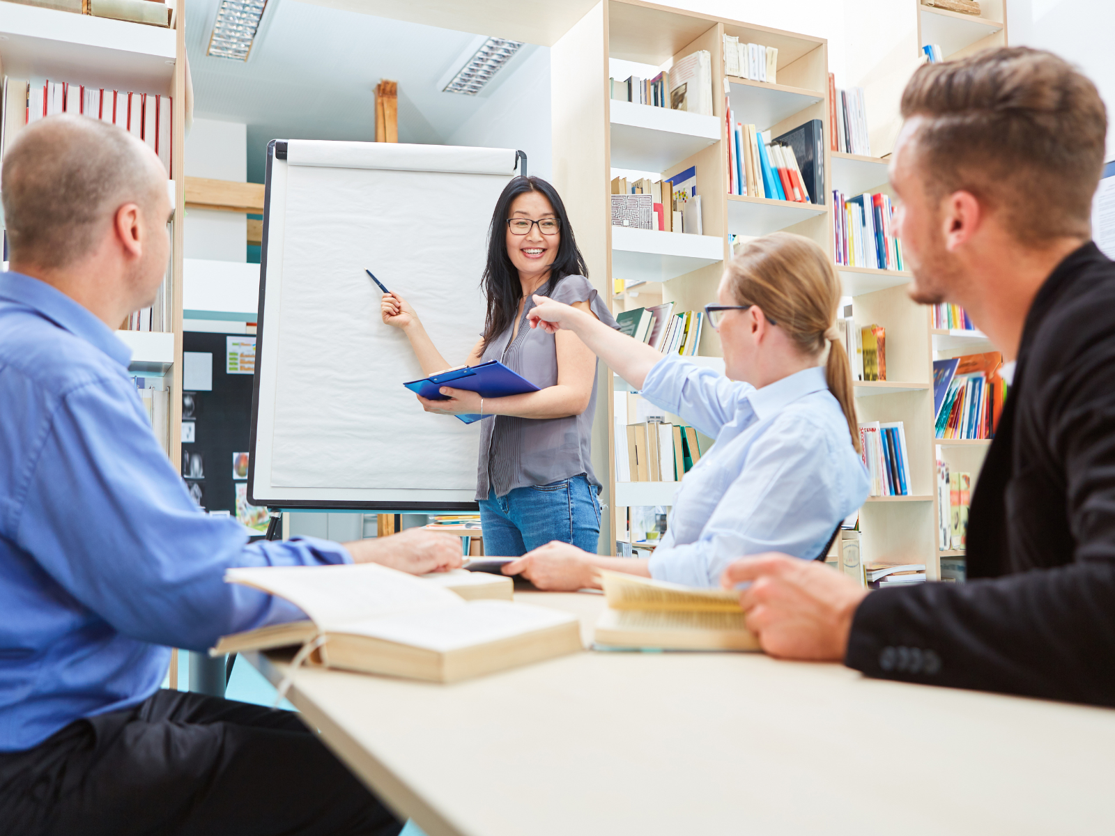 a woman standing in front of a whiteboard with a pen and a group of people sitting around