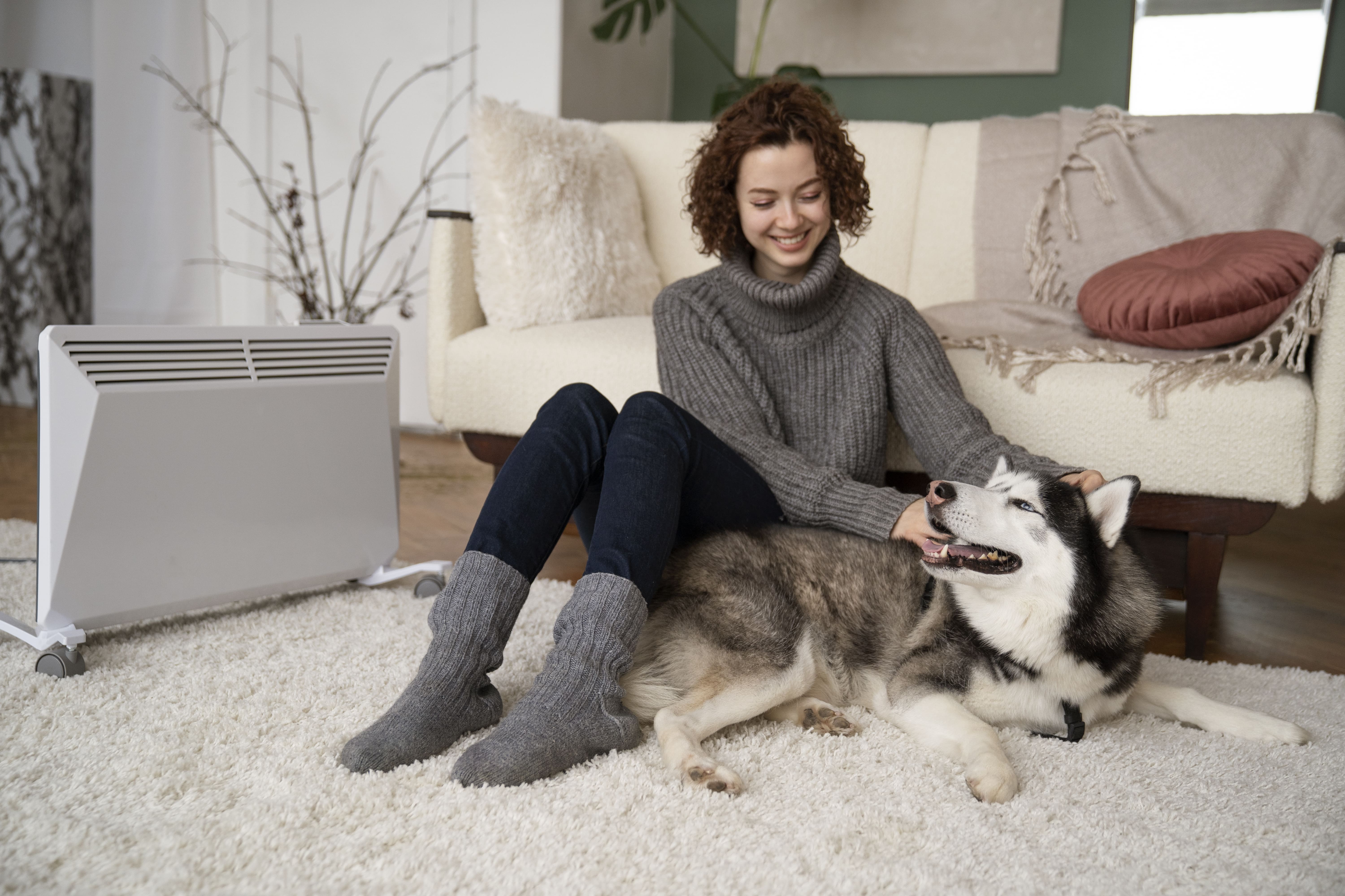 Smiling woman in cozy sweater petting husky dog by electric heater