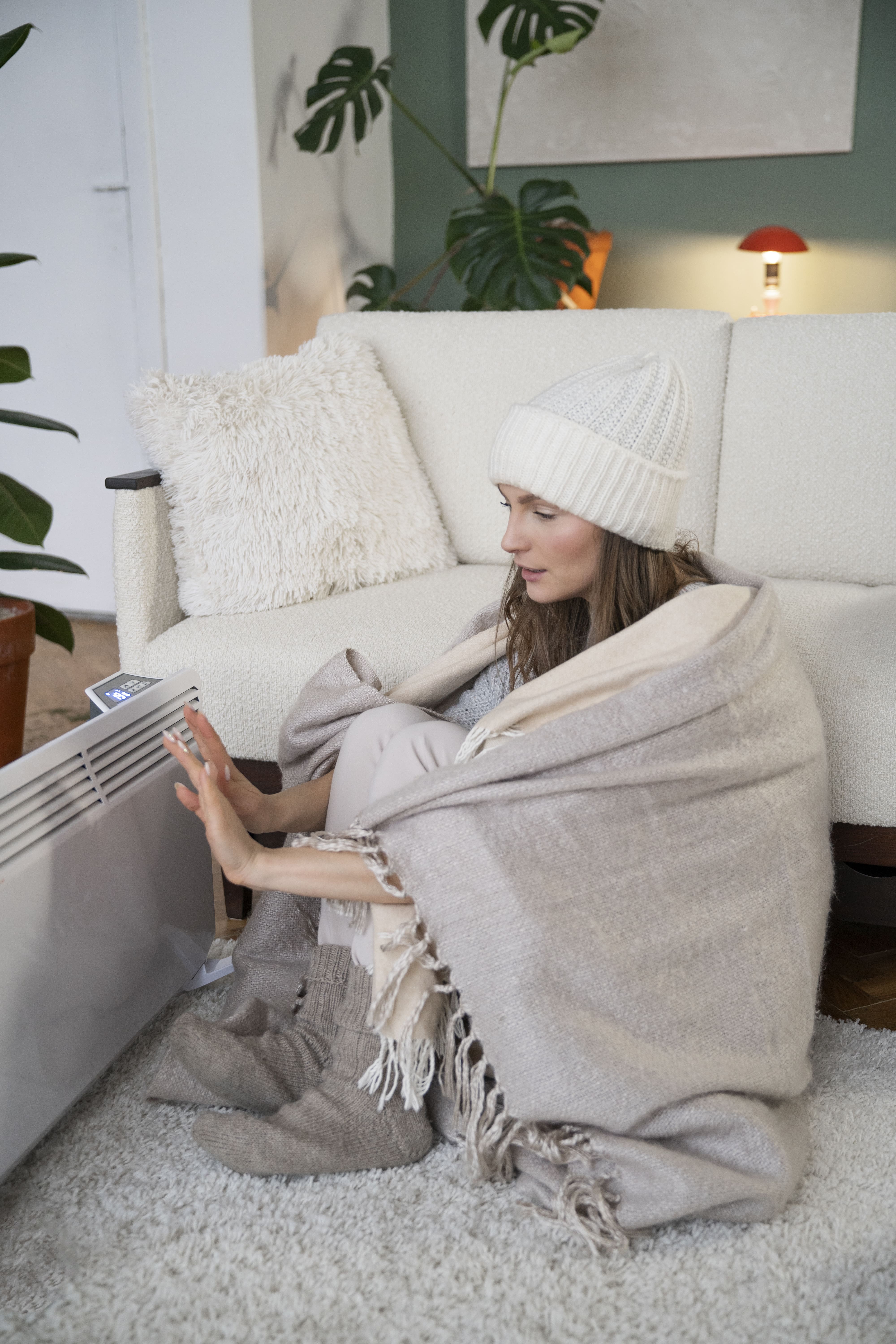 Cozy person in white beanie adjusting heater while wrapped in blanket