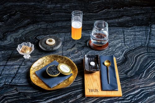 Elegant table setting with a golden plate holding a tin of black caviar, small dish with a white pill, gold spoon, a glass of clear water on a wooden coaster, and a flute of orange cocktail on a dark textured surface.