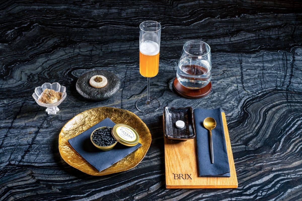 Elegant table setting with a golden plate holding a tin of black caviar, small dish with a white pill, gold spoon, a glass of clear water on a wooden coaster, and a flute of orange cocktail on a dark textured surface.
