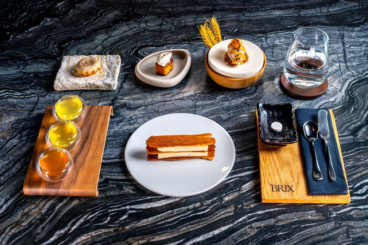 Elegant dessert presentation including layered pastry on white plate, three small glass cups with colorful liquids on a wooden board, a glass of water, a napkin with cutlery, and smaller plated desserts on a dark textured table.