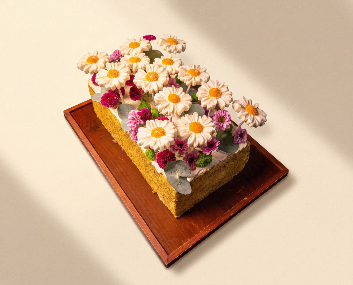 Rectangular pistachio cake topped with white chocolate daisies and fresh flowers on a wooden tray