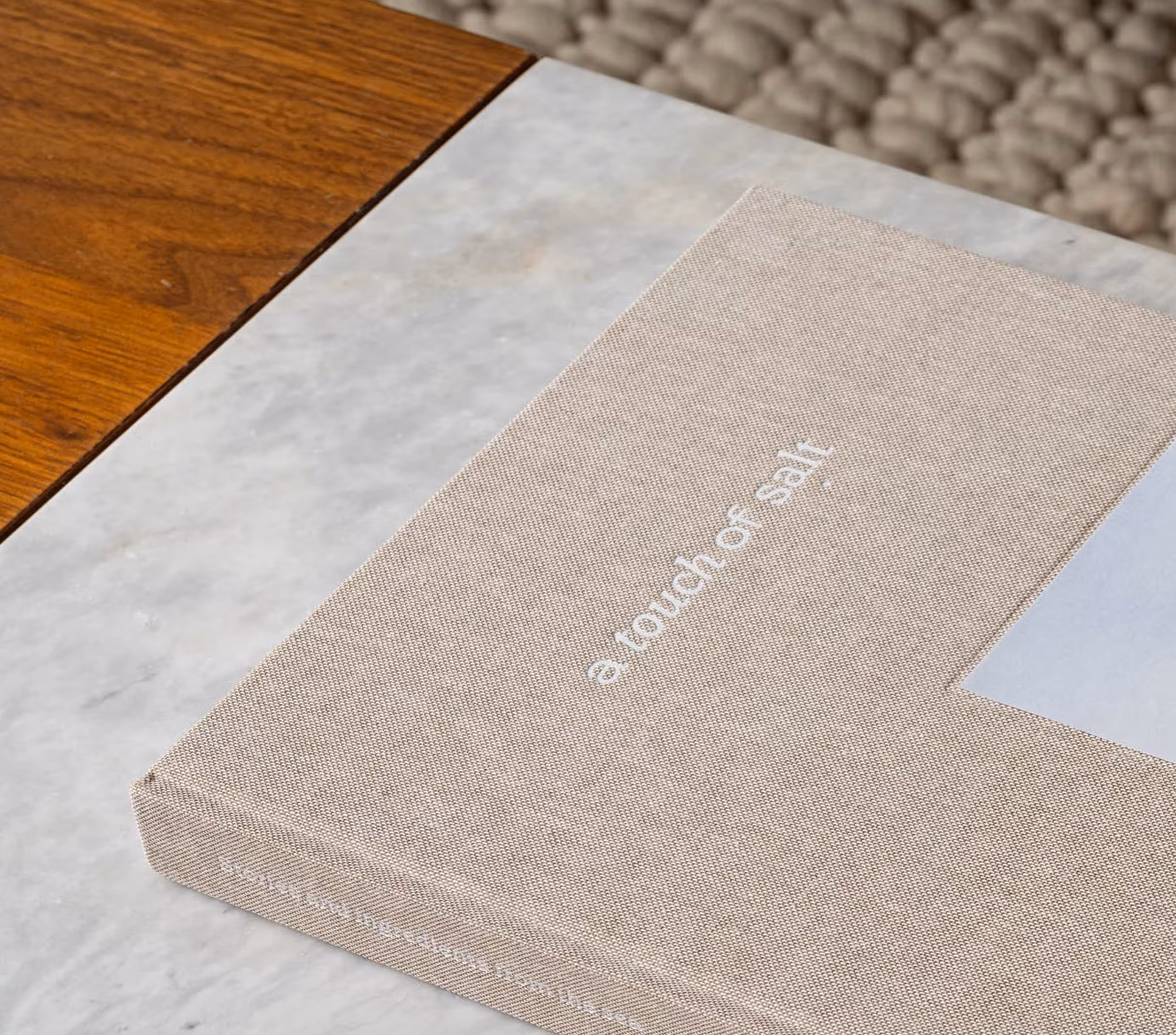 Beige cloth-covered book titled 'a touch of salt' placed on a marble surface next to a wooden table.