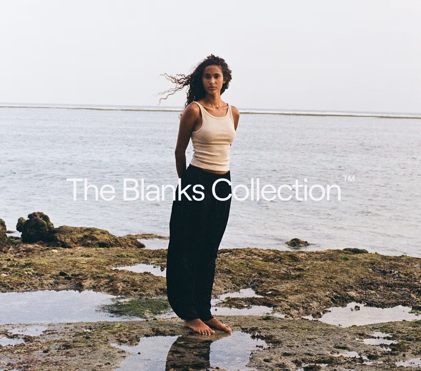 A woman with curly hair wearing a beige tank top and black pants stands barefoot on rocky ground near the water's edge.