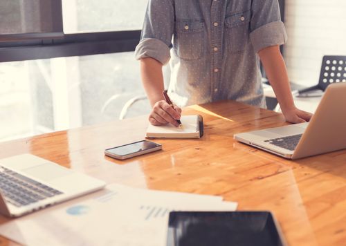 Person in a denim shirt writing in a notebook on a wooden desk with two laptops, a smartphone, and papers scattered around.