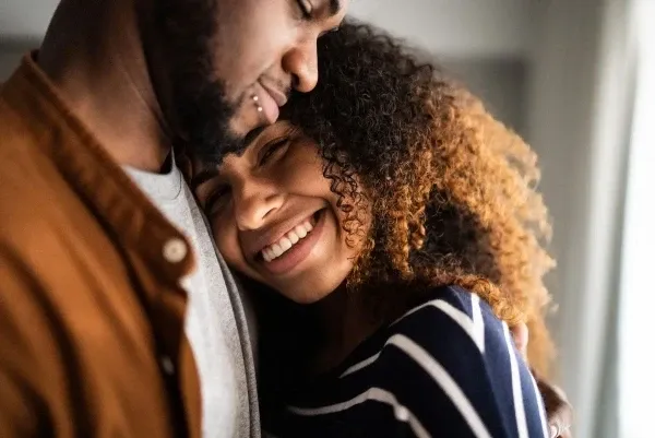 A smiling woman with curly hair hugging a man with a beard and lip piercing.