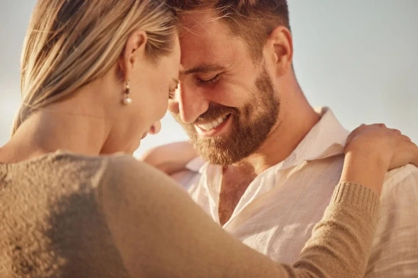 Close-up of a smiling couple embracing with foreheads touching outdoors in soft sunlight.