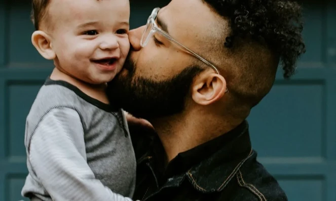 Man with glasses kissing a smiling baby on the cheek in front of a blue door.