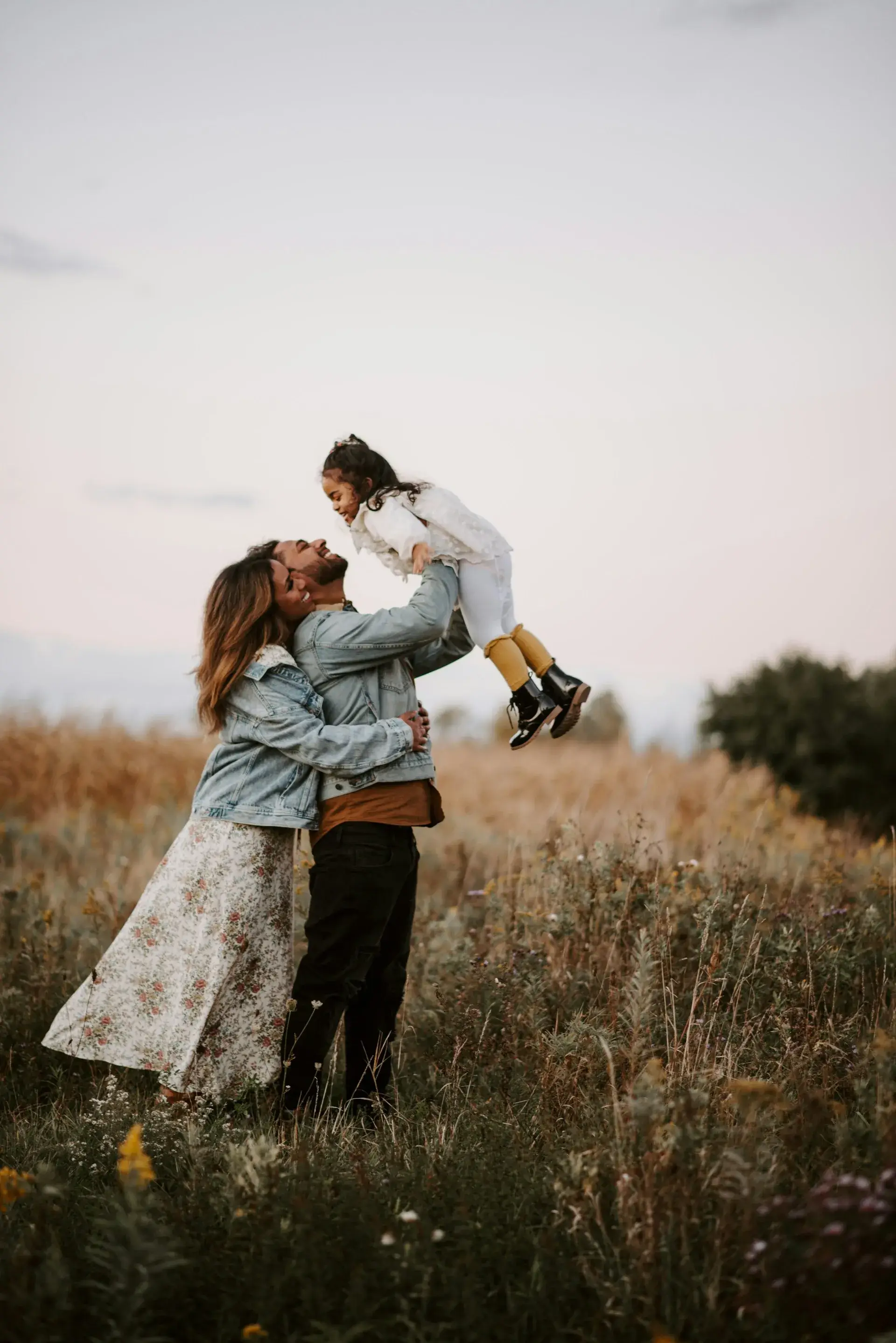 A man and woman stand in a field of tall grass at sunset, smiling and holding a young girl up in the air.