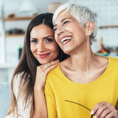 Two women smiling and embracing indoors, one with short gray hair wearing yellow and the other with long dark hair wearing white.