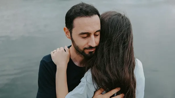Man and woman embracing near a body of water, with the man closing his eyes gently.