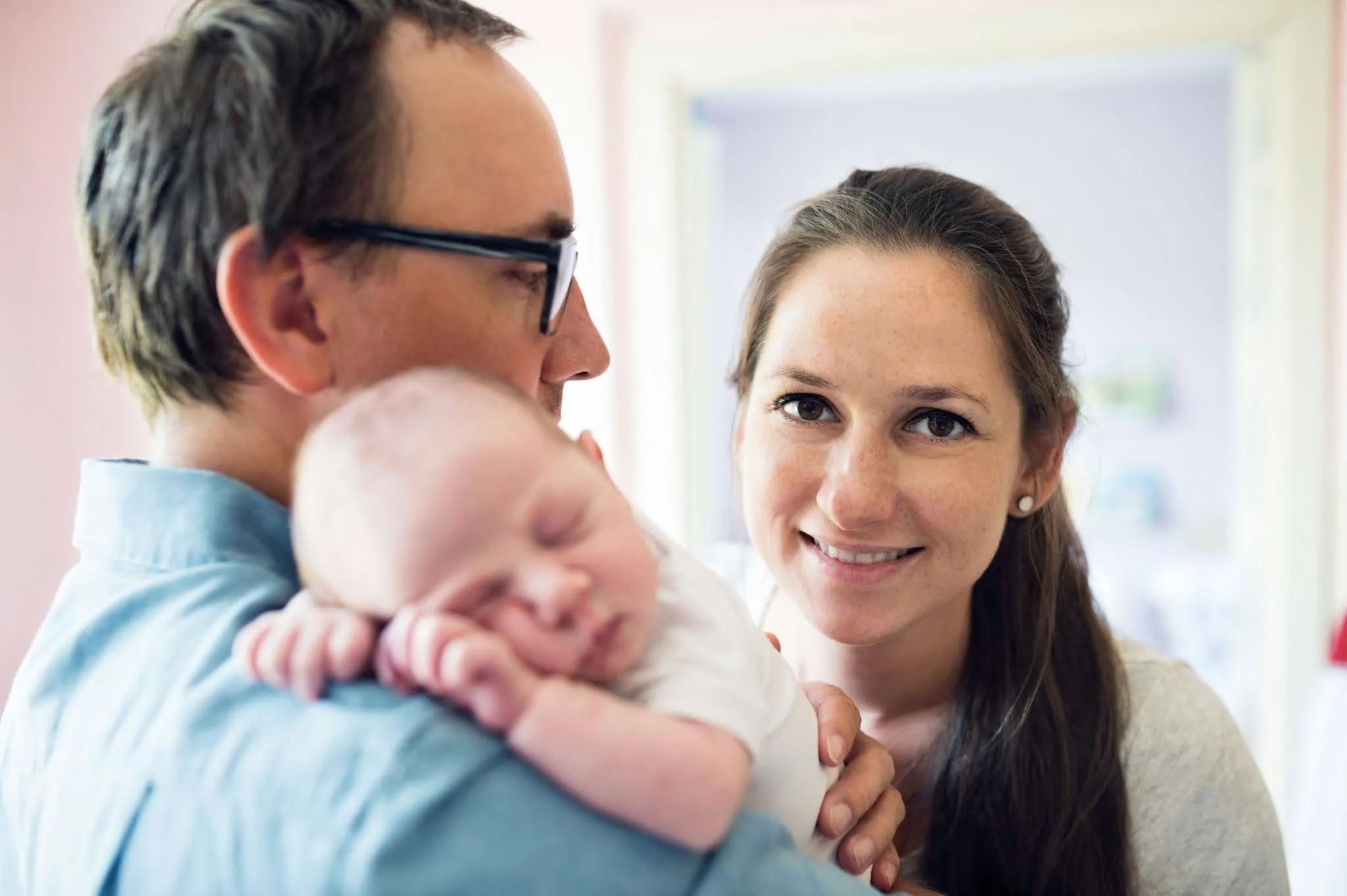 Smiling woman looking at camera as man holds a sleeping baby on his shoulder indoors.