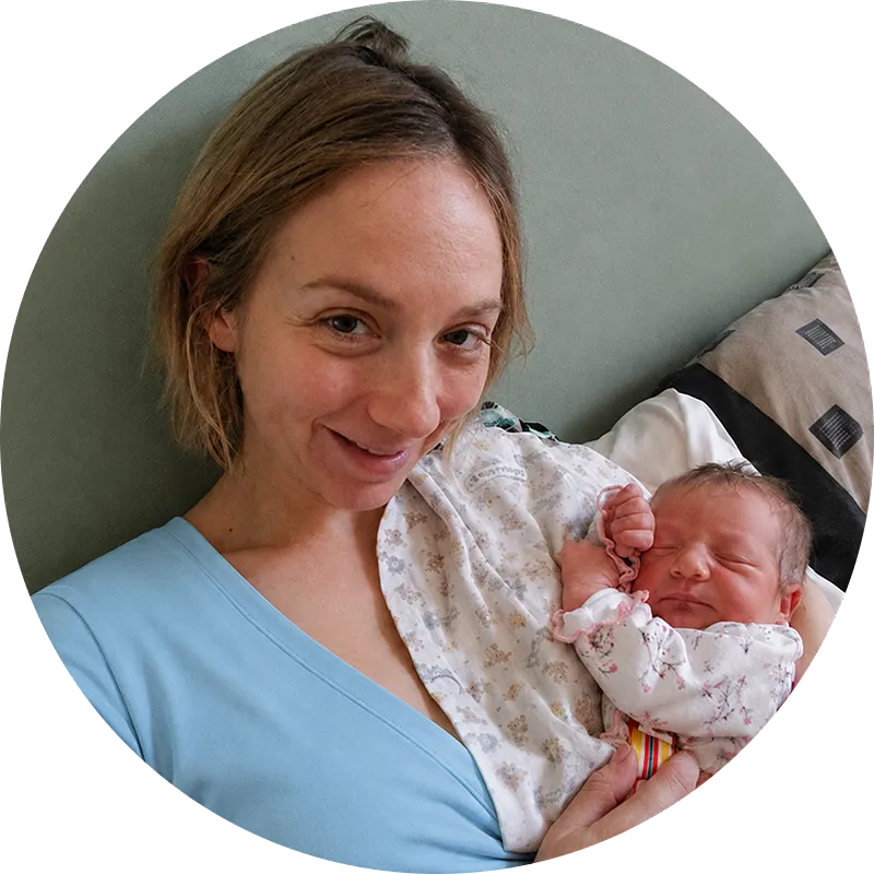 Woman smiling while holding a newborn baby wrapped in a white blanket with a patterned pillow behind.