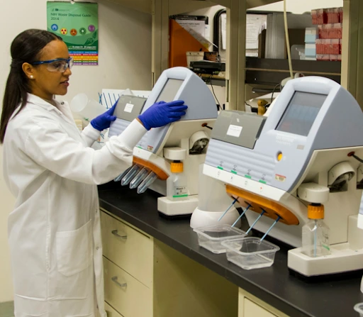 Scientist in lab coat and gloves operating gene testing machines in a laboratory setting.