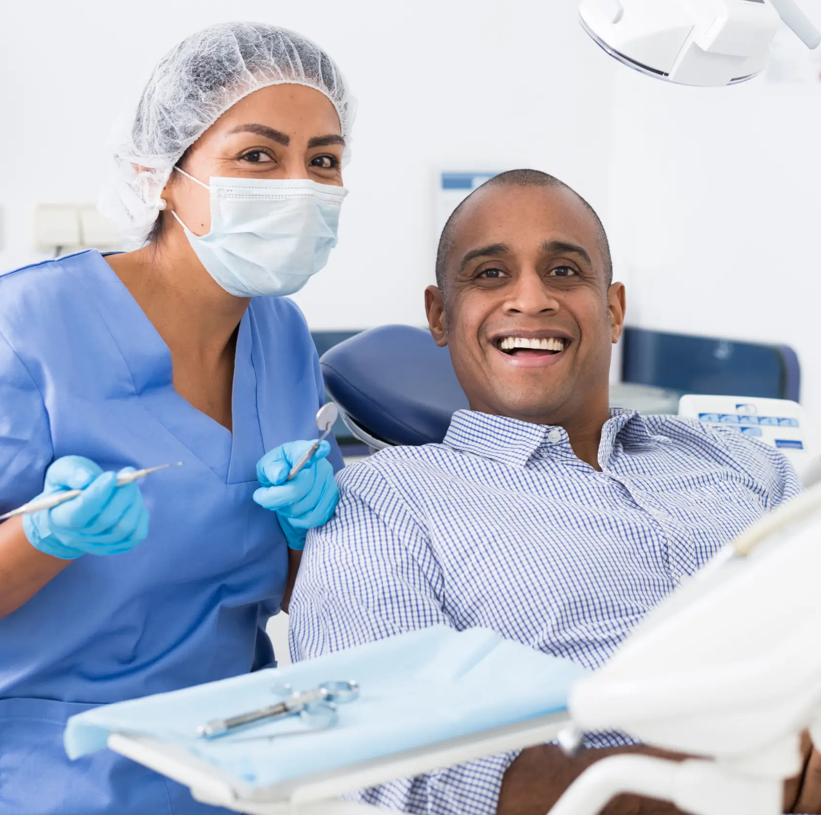 A dentist in blue scrubs and mask stands beside a smiling patient in a dental chair.