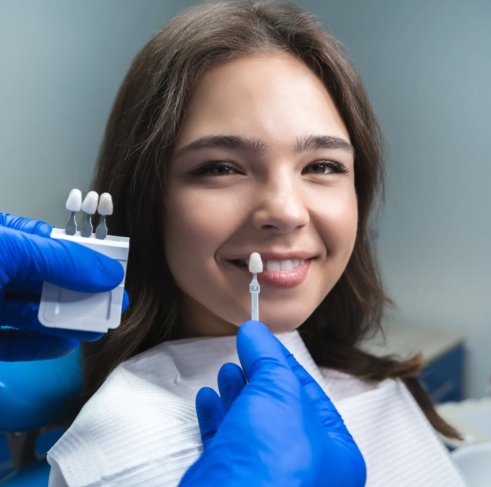 A dentist matches tooth shade samples to a smiling woman's teeth in a dental clinic.
