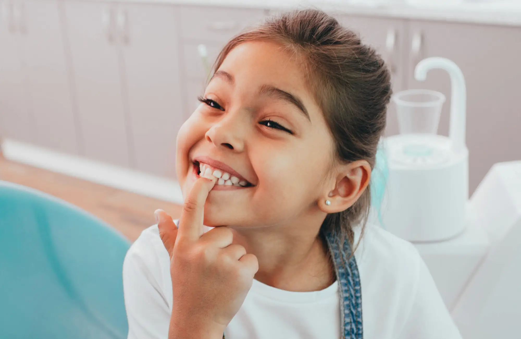 A young girl sitting in a dental chair smiles and points to her teeth.