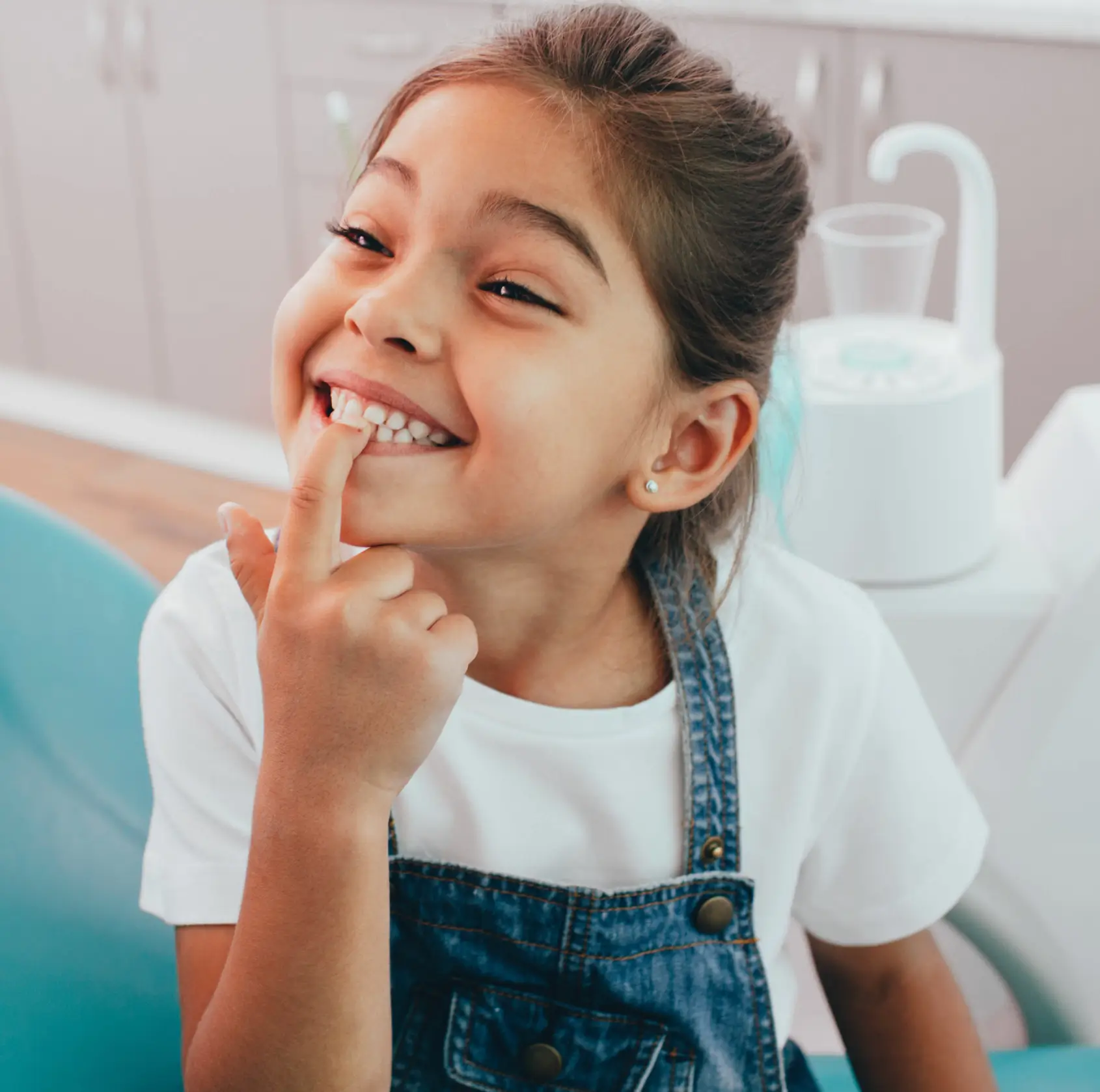 A young girl in denim overalls points to her missing tooth and smiles at the dentist's office.