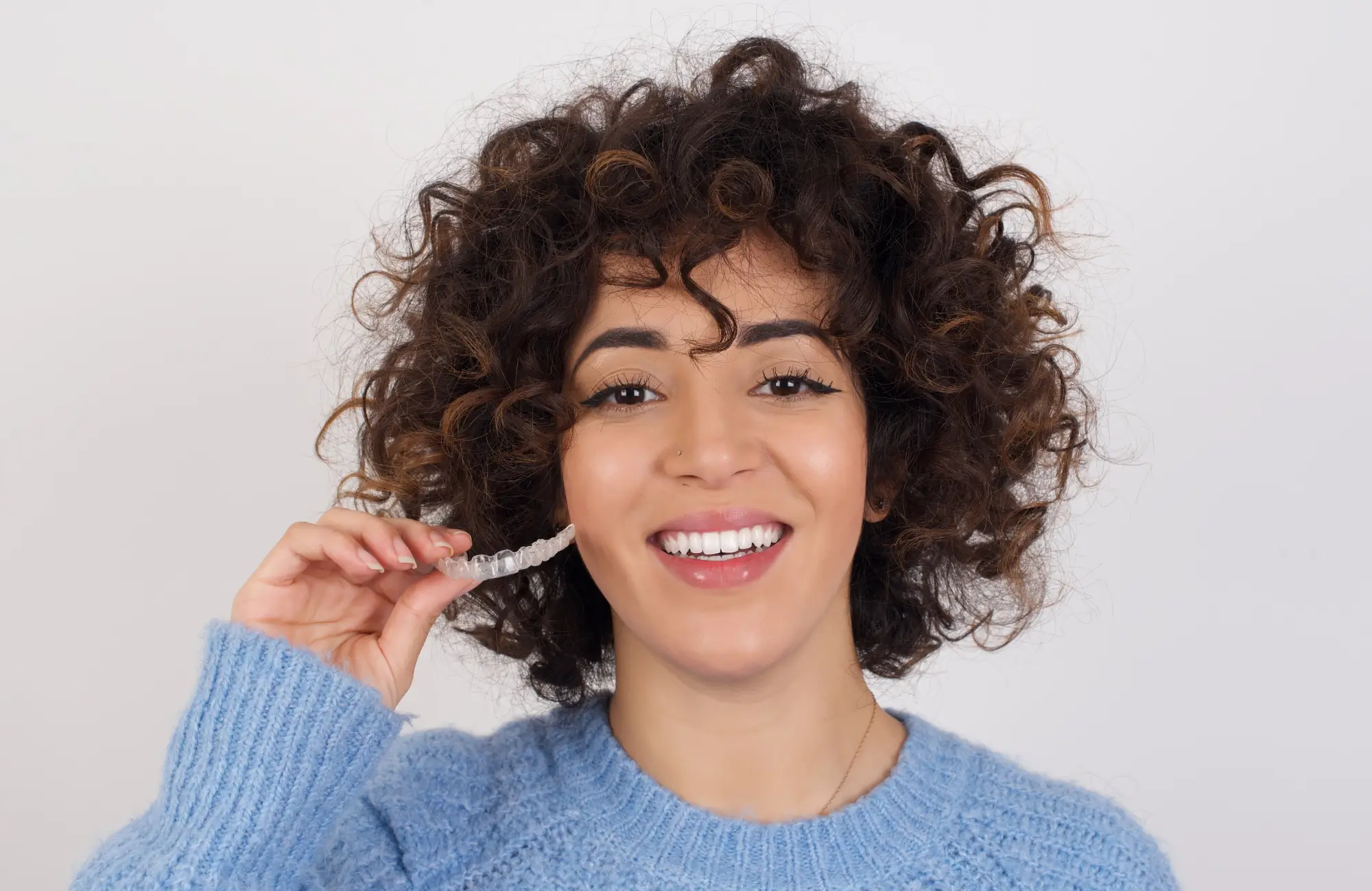 Person smiling while holding a clear dental aligner near their teeth.
