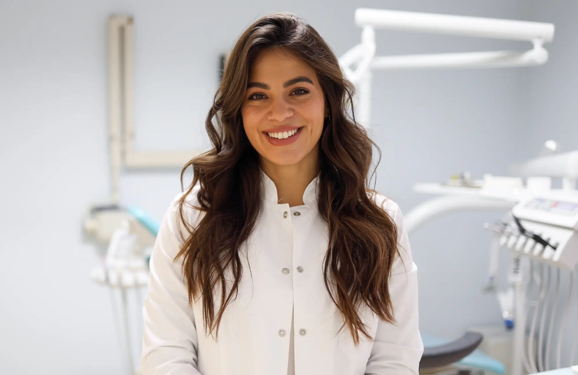 A woman wearing a white coat smiles, standing in a dental office.