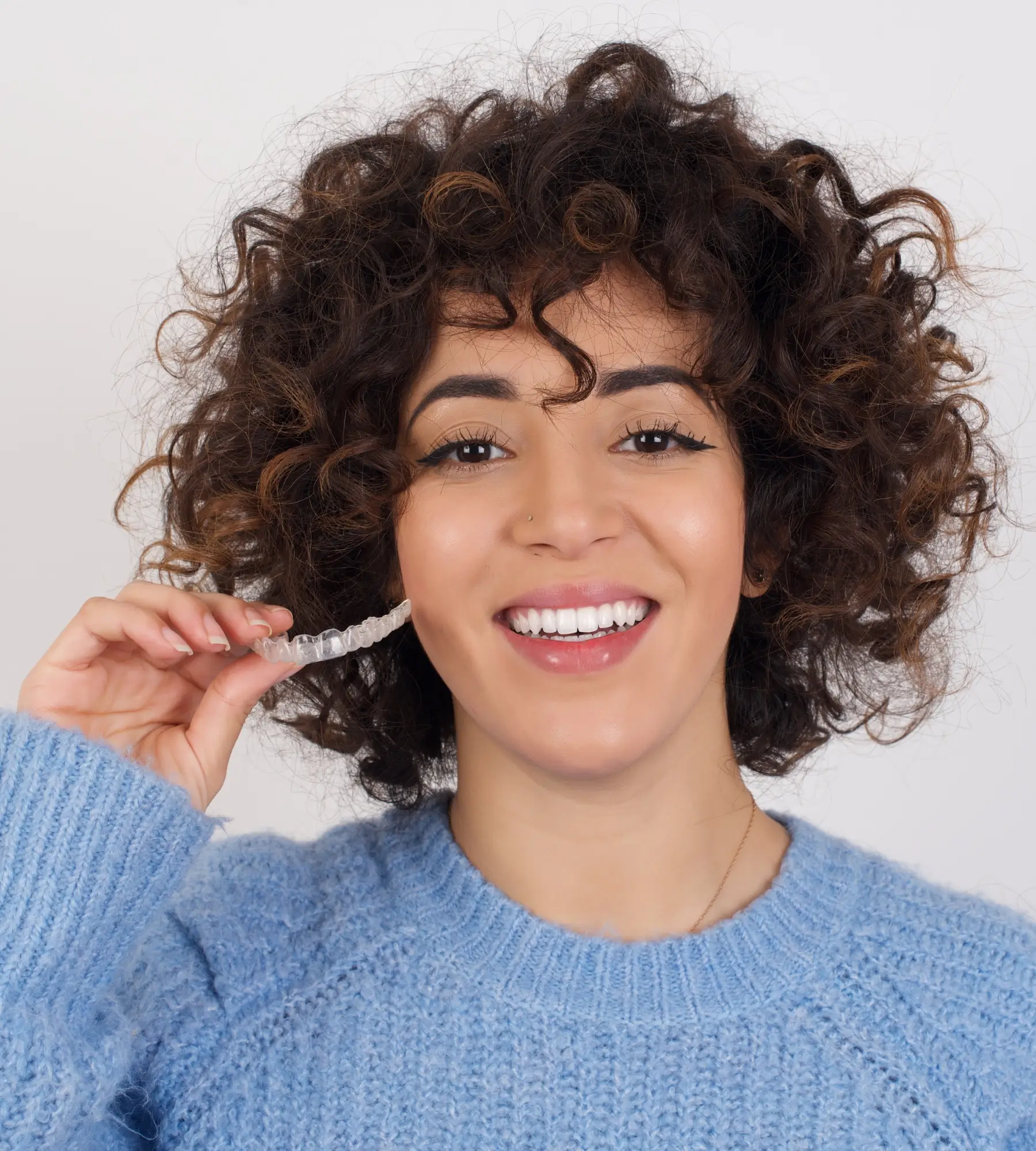 A woman in a blue sweater smiles while holding a clear dental aligner near her mouth.
