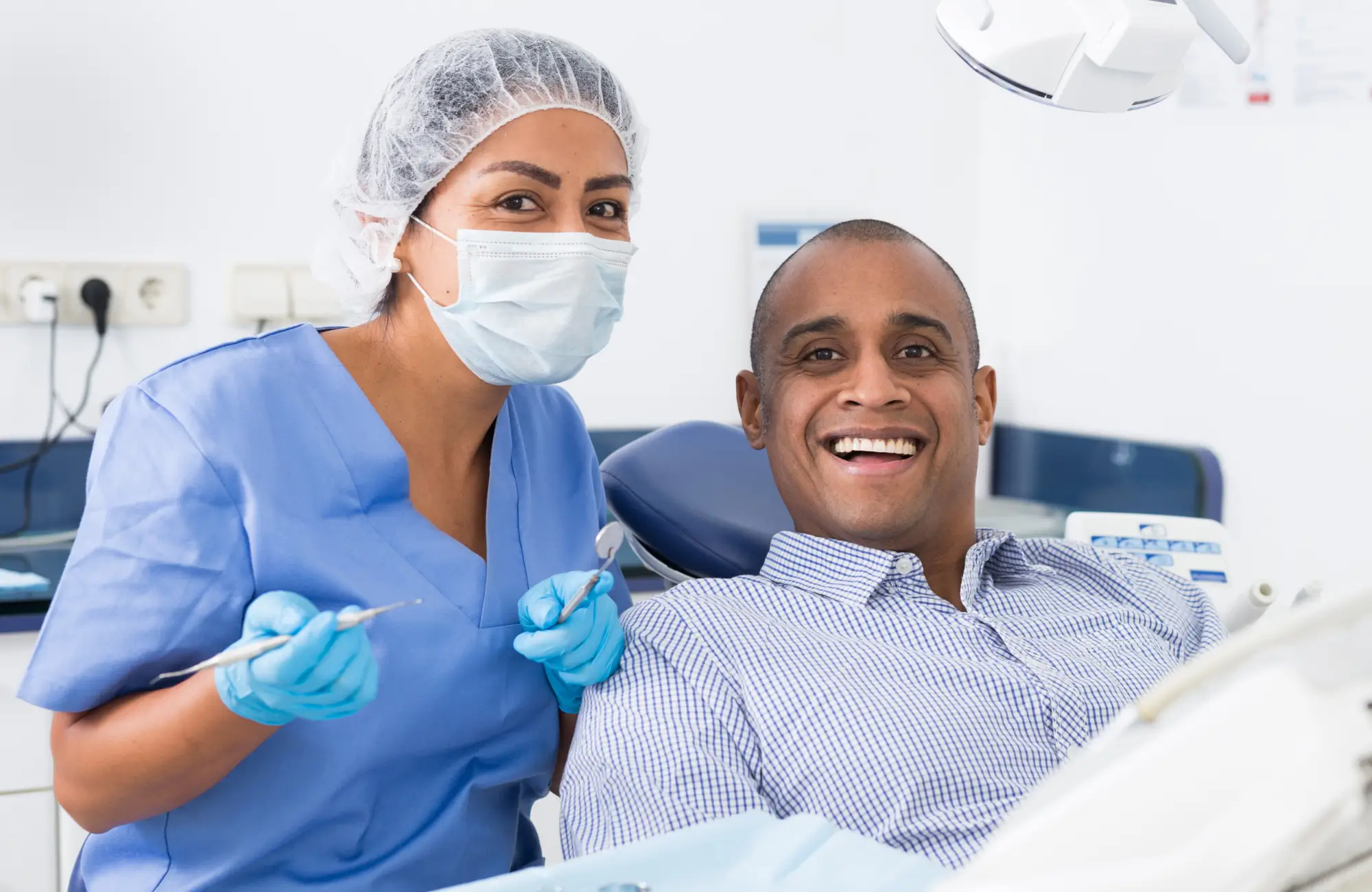 A dentist smiles behind a mask while a patient sits in a chair, also smiling.