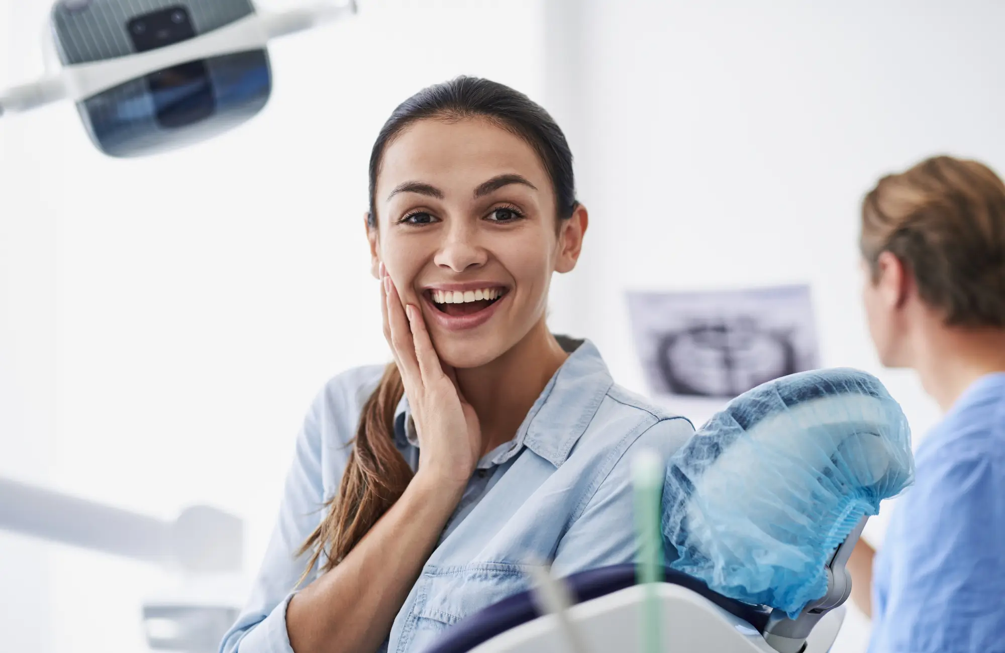 Woman in dental office smiling with hand on cheek, dental chair and equipment in the foreground.