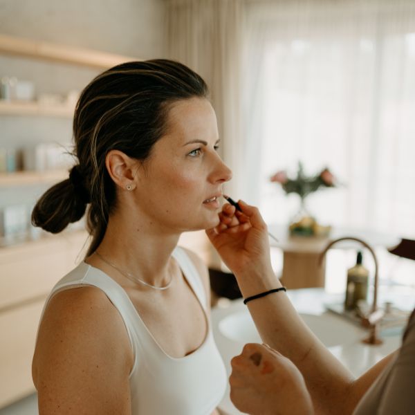 Žena ve světlém tílku si nechává nanášet rtěnku v osvětlené místnosti.
